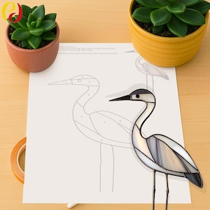 May include: A stained glass heron with a black beak and a gray and white body. The heron is standing on a piece of paper with a heron outline and numbers. Two potted succulents are on either side.