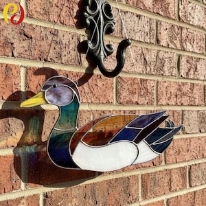 Könnte beinhalten: Glasmalerei-Ente mit gelbem Schnabel und farbenfrohem Körper in Blau-, Grün-, Braun- und Weißtönen. Die Ente hängt an einer Backsteinmauer. Die Ente ist ein dekorativer Gegenstand.