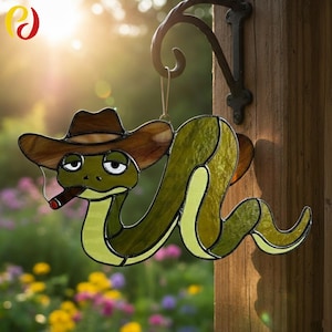 May include: A stained glass snake ornament, depicted with a brown cowboy hat and a cigar. The snake is green and yellow with black outlines, suspended from a black metal hook. The background shows a blurred garden.