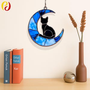 Peut inclure: Une fenêtre en verre coloré suspendue représente un chat noir assis sur une lune en croissant bleue. La lune a un motif en verre coloré blanc et bleu.