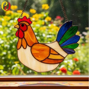May include: Stained glass rooster suncatcher with a red comb, orange body, and blue and green tail feathers. The decorative rooster is hanging from a chain. The background is a blurred view of a garden.