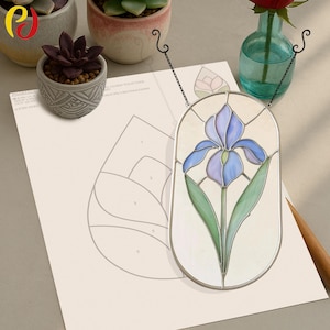 May include: Stained glass iris window hanging with a silver metal frame. The iris features blue and purple petals with green leaves. A paper with a floral design is in the background. Small potted succulents and a vase with a red rose are also visible.