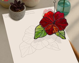 May include: A red stained glass hibiscus flower with green leaves, suspended by a chain. The flower rests on a paper with a flower outline and numbers. Small potted plants and a rose in a vase are in the background.