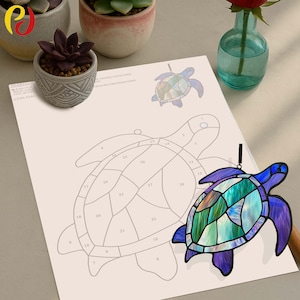 Op de afbeelding: Een gebrandschilderde zonnevanger in de vorm van een schildpad met een blauw, groen en paars ontwerp. De schildpad hangt naast een papier met een schildpadomtrek en genummerde secties. De afbeelding bevat ook kleine potplanten en een vaas met een rode roos.