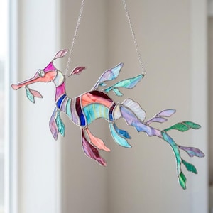 May include: A stained glass suncatcher shaped like a leafy seadragon, featuring multicoloured glass panels in shades of pink, blue, purple, and orange, held together by silver metal solder lines and suspended by a thin silver chain for display.