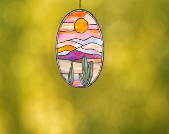 May include: A stained glass window hanging with a sunset scene over a desert landscape. The scene features a pink and orange sky, purple mountains, and green cacti.