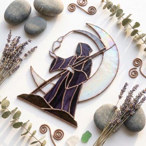 Peut inclure: Un attrape-soleil en vitrail représentant une silhouette encapuchonnée vêtue d'une robe violet foncé tenant une longue faux, placée devant un grand croissant de lune irisé. L'objet est assemblé avec des lignes de soudure en métal sombre.