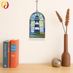 Puede incluir: Vidriera de faro con marco de metal negro. El faro presenta rayas azules y blancas, sobre un cielo azul y un paisaje verde. Colgando de una cadena negra. Elementos decorativos: libros, un jarrón y un huevo plateado.