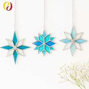 May include: Three star-shaped stained glass ornaments in shades of blue and white, hanging from silver chains. Each star is outlined in silver. The ornaments are displayed against a white background.