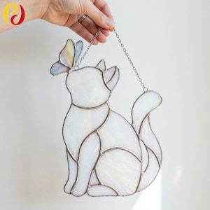 May include: A stained glass cat figurine with a butterfly. The cat is white with a silver outline and a chain for hanging. The butterfly has pastel colors. A decorative item for cat lovers.
