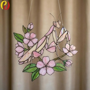 May include: A stained glass suncatcher featuring a pink praying mantis perched among light pink cherry blossoms and green leaves. The piece is constructed with soldered metal lines and iridescent glass segments, suspended by thin clear filaments.