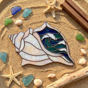 May include: A stained glass conch shell ornament featuring iridescent white glass and a central section of deep blue and teal glass depicting a wave. The piece is surrounded by scattered seashells, sea glass, and a starfish on sand.