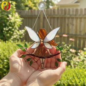May include: Stained glass fairy suncatcher with iridescent wings, brown hair, and a brown dress. The fairy is perched on a branch with green leaves. It hangs from a silver chain and suction cup, perfect for window display.