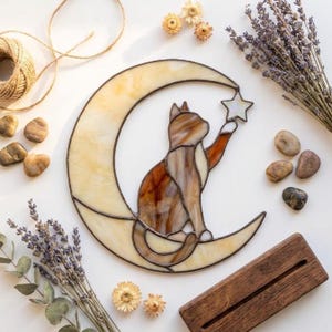 Peut inclure: Un attrape-soleil en vitrail représentant un chat brun et crème assis sur un croissant de lune jaune pâle, tendant la patte vers une petite étoile blanche. L'objet est assemblé avec des lignes de soudure en métal sombre et fini lisse.