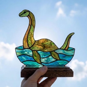 May include: A stained glass sculpture of a long-necked creature resembling a plesiosaur emerging from blue and teal waves. The creature is crafted from textured green and amber glass pieces, all mounted on a rectangular wooden base.