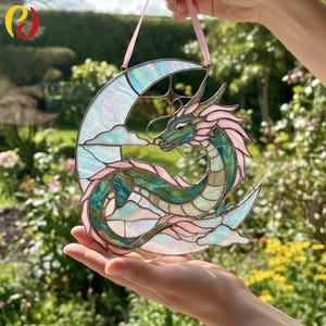 May include: Stained glass dragon and moon hanging decoration. The dragon is green and pink, with a crescent moon and clouds. The decoration is held by a pink ribbon. The background is a blurred view of a garden.