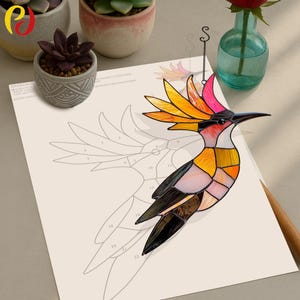 May include: A stained glass hummingbird with a colourful design, including shades of pink, orange, yellow, and black. The hummingbird is hanging from a silver chain and is positioned over a numbered template for a stained glass project.