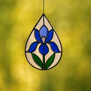 Könnte beinhalten: Ein tropfenförmiger Glas-Suncatcher mit einem blauen Irisblumen-Design. Die Blume hat blaue Blütenblätter, eine lavendelfarbene Mitte und grüne Blätter. Das Glas ist mit schwarzen Linien umrandet und hängt an einer Kette.