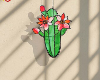 May include: A stained glass cactus with vibrant green body and red and pink flowers. The cactus is suspended by a chain, casting a shadow on the beige wall. The glass art piece is a decorative item.