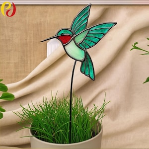 May include: A stained glass hummingbird garden stake with a black metal stem. The hummingbird features green wings, a red throat, and a white belly. The stake is placed in a pot of green grass, creating a decorative garden accent.