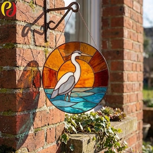 May include: A round stained glass panel depicting a heron standing in water, with a sunset background. The panel is in shades of orange, blue, and white, and is hanging from a decorative metal bracket. The panel is approximately 10 inches in diameter.