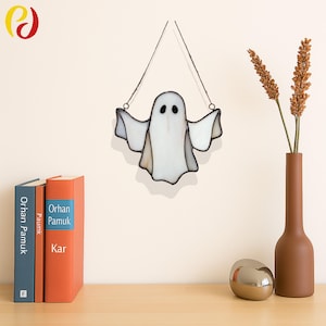 May include: A white and clear stained glass ghost decoration with black eyes and outstretched arms, hanging from a thin wire. The Halloween-themed decor is displayed against a neutral wall, with a vase of dried flowers and books on a wooden surface.