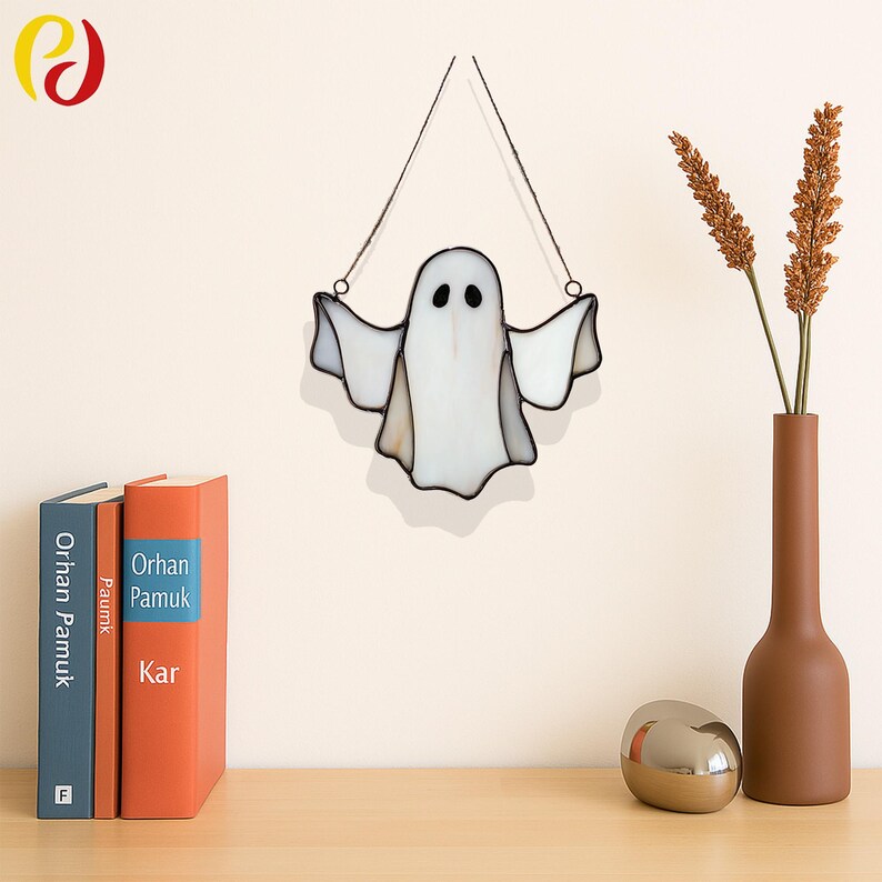May include: A white and clear stained glass ghost decoration with black eyes and outstretched arms, hanging from a thin wire. The Halloween-themed decor is displayed against a neutral wall, with a vase of dried flowers and books on a wooden surface.