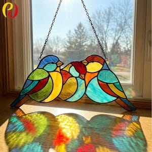 May include: A stained glass window decoration depicting three birds in vibrant colours. The birds are made of blue, green, yellow, orange, and red glass. The window is suspended by a chain and hangs in front of a window with a view of trees.