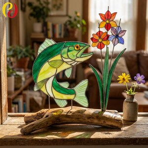 May include: A stained glass fish sculpture in green and yellow, displayed on driftwood. Next to it are colourful glass flowers in red, orange, and purple. A small vase with yellow and purple flowers is also present.