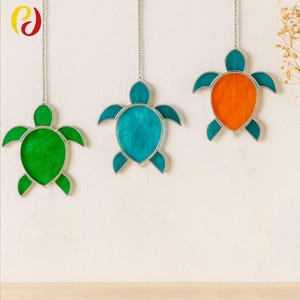 Puede incluir: Tres adornos de tortuga de vidrieras en verde, turquesa y naranja, cada uno con un marco de metal plateado y una cadena para colgar. Las tortugas están suspendidas contra una pared blanca, creando una exhibición decorativa.