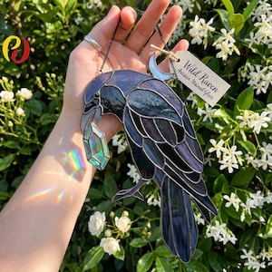 May include: Stained glass raven suncatcher with a gemstone in its beak. The raven is primarily dark blue and purple, with a silver chain for hanging. The tag reads "Wild Raven Artisan Stained Glass".