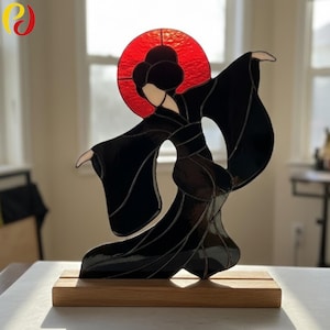May include: Stained glass sculpture of a woman in a black kimono with a red sun behind her head. The figure is mounted on a rectangular wooden base. The woman's face and hands are white, and the sun is a vibrant red.