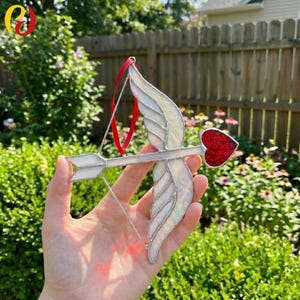 May include: A stained glass Cupid's arrow with a red heart-shaped arrowhead and white feathered wings. The arrow is held by a hand against a backdrop of green foliage and a wooden fence. A red ribbon is attached for hanging.