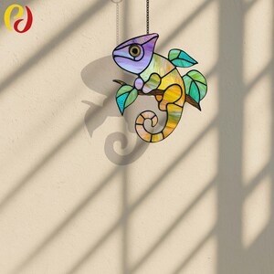 May include: A colorful stained glass chameleon suncatcher hanging from a chain. The chameleon is perched on a branch, with a vibrant mix of purple, yellow, green, and blue hues. The suncatcher casts a shadow on the wall.