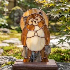Puede incluir: Una figura de vidriera que representa un tanuki, o perro mapache japonés, hecha con piezas de vidrio en tonos marrón y crema unidas por cobre. La figura está montada sobre una base de madera rectangular y tiene un cuerpo redondeado.