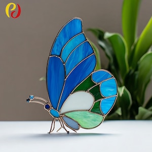 May include: A stained glass butterfly sculpture with blue, green, and white panels. The butterfly has a copper-colored frame and delicate antennae. The wings feature various shades of blue and green, with a white center. Decorative art piece.