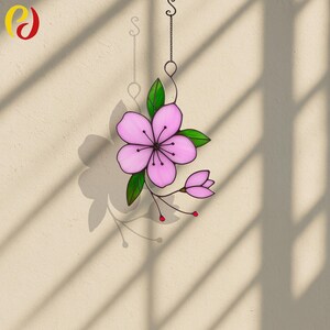 May include: A decorative stained glass flower in shades of pink and green, suspended by a metal chain. The flower features a central bloom and a bud. The background is a beige wall with shadows.