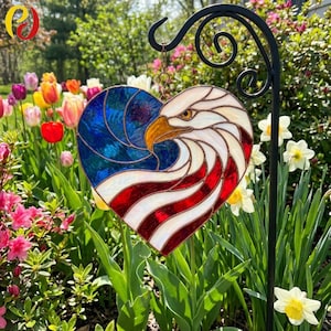 May include: A heart-shaped stained glass suncatcher featuring a bald eagle head design integrated with the American flag. The piece displays blue, white, and red glass segments with a yellow beak, all held together by dark metal solder lines.