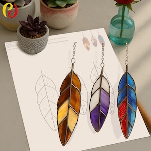Op de afbeelding: Drie gebrandschilderde veervormige ornamenten in bruin-, paars- en blauwtinten, elk opgehangen aan een ketting. De veren worden weergegeven op een vel papier met een bladpatroon en genummerde secties. Decoratieve vetplanten en een vaas staan op de achtergrond.