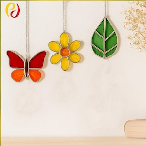 May include: Three stained glass ornaments hanging from chains. A butterfly with red and orange wings, a yellow flower with an orange center, and a green leaf. The ornaments are set against a neutral background.