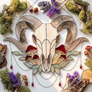 May include: A stained glass wall hanging featuring a ram skull in cream and brown tones, surrounded by red mushrooms and pale green leaves. The piece is accented with silver chains and small red glass teardrop charms hanging from the bottom.