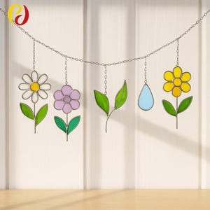 May include: A decorative hanging featuring stained glass flowers, leaves, and a teardrop. The flowers are in yellow, purple, and white with green leaves. The teardrop is light blue. All pieces are connected by a silver chain.