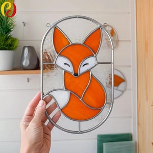 Puede incluir: Vidriera con un zorro. El zorro es de color naranja y blanco, con un marco de metal plateado. El diseño tiene forma ovalada. El fondo es de vidrio transparente. La obra de arte está colgada en una pared blanca.