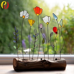 Könnte beinhalten: Eine dekorative Blumenskulptur mit bunten Glasmalereiblüten und -blättern. Die Blumen sind in Weiß, Orange, Gelb und Rot gehalten, mit grünen und blauen Blättern. Die Stiele sind aus schwarzem Draht, und die Basis ist ein Stück Treibholz.