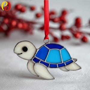 Könnte beinhalten: Ein Schildkröten-Ornament aus Glasmalerei mit einem roten Band zum Aufhängen. Die Schildkröte hat einen weißen Kopf und Flossen, einen blauen und hellblauen Panzer und ein schwarzes Auge. Das Ornament ist mit einer silbernen Metallkontur versehen.