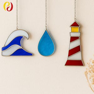 May include: Three stained glass ornaments hanging from chains. The ornaments include a blue and white wave, a blue teardrop, and a red and white striped lighthouse with a yellow top. The ornaments have silver metal borders.