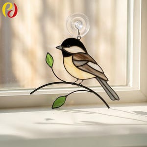 May include: Stained glass bird suncatcher featuring a chickadee perched on a black metal branch with green leaves. The bird has a black head, tan body, and brown and white wings. A suction cup is attached for window display.