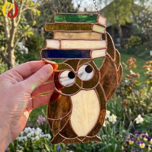 Puede incluir: Un búho de vidriera sosteniendo una pila de libros. El búho es de vidrio marrón, blanco y ámbar, con ojos grandes. Los libros son en tonos verdes, amarillos y azules. La pieza está diseñada para colgarse y está enmarcada con cobre.