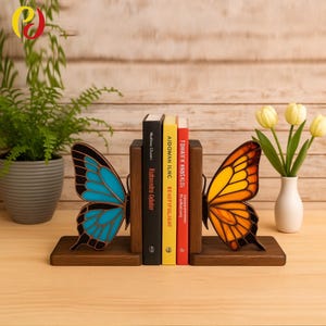 Könnte beinhalten: Buchstützen aus Holz in Schmetterlingsform mit dunkelbraunem Sockel und bunten Flügeln. Ein Flügel ist blau und der andere orange und gelb. Drei Bücher stehen zwischen den Buchstützen. Ein Farn und Tulpen sind im Hintergrund.