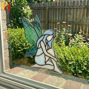 May include: A stained glass fairy with blue hair and iridescent green wings. The fairy is in a seated position, with its arms wrapped around its knees. The glass is set in a window, with a green garden visible in the background.